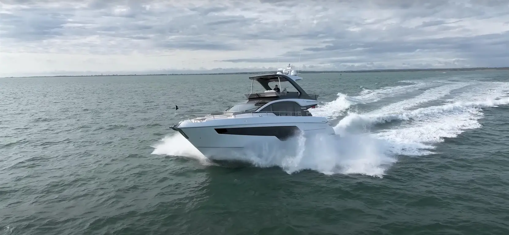 A luxury motor yacht speeds through the ocean, creating white spray against a cloudy sky.